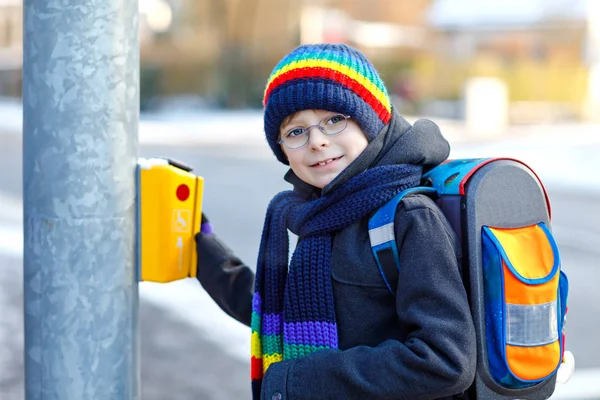 Kleiner Schulbub der Grundschule, der bei Schneefall zur Schule läuft. Glückliches gesundes Kind mit Brille auf Knopf für Ampel. Mit Rucksack oder Schulranzen in bunter Winterkleidung. — Stockfoto