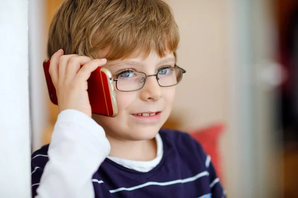 Милый маленький мальчик в очках говорит по сотовому телефону. Очаровательный здоровый ребенок держит смартфон и разговаривает с подругой или с родителями, бабушкой и дедушкой. — стоковое фото