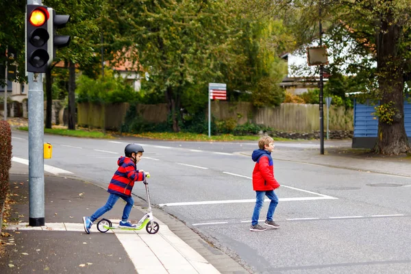 Two little schoolkids boys running and driving on scooter on autumn day. Happy children in colorful clothes and city traffic crossing pedestrian crosswalk with traffic lights. — Stock Photo, Image