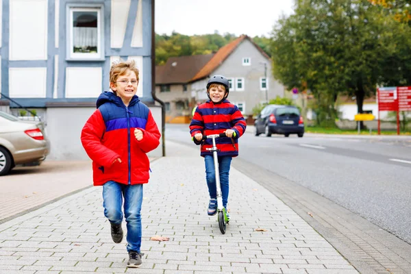 Dwóch małych chłopców schoolkids bieganie i jazda na skuterze na jesienny dzień. Szczęśliwe dzieci w ruchu miejskim i kolorowe ubrania. Rodzeństwo, zabawy na świeżym powietrzu, aktywny wypoczynek dla dzieci — Zdjęcie stockowe