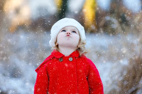 Odkryty zimowy portret małej uroczej dziewczynki w czerwonym płaszczu i białym kapeluszu. Zdrowe, szczęśliwe dziecko spacerujące po parku w chłodny dzień ze śniegiem i opadami śniegu. Stylowe ubrania dla dzieci. — Zdjęcie stockowe