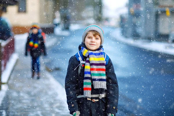 Dos niños pequeños de clase primaria caminando a la escuela durante las nevadas. Niños felices divirtiéndose y jugando con la primera nieve. Hermanos y amigos con mochila en ropa de invierno colorida. — Foto de Stock