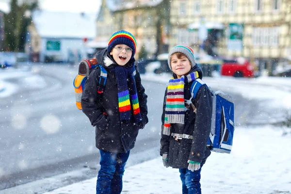 Dwójka małych chłopców z podstawówki, idących do szkoły podczas opadów śniegu. Szczęśliwe dzieci bawiące się i bawiące z pierwszym śniegiem. Rodzeństwo przyjaciele z plecakiem w kolorowe ubrania zimowe. — Zdjęcie stockowe