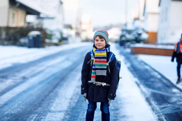 Malý školáček základní třídy, který chodí do školy během sněžení. Šťastné dítě baví a hrát si s prvním sněhem. Student s batohem nebo brašnou v barevných zimních šatech. — Stock fotografie