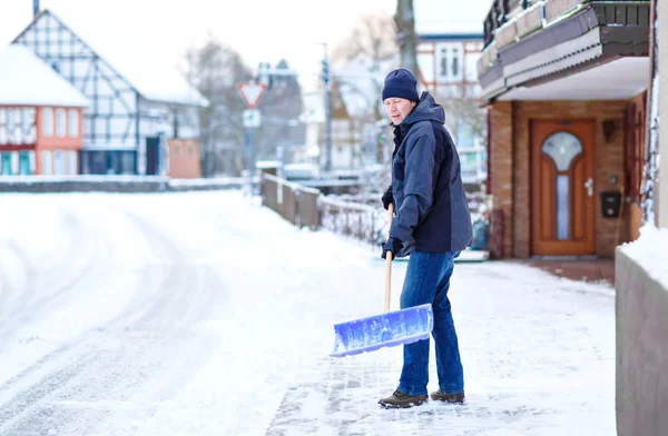 Man with snow shovel cleans sidewalks in winter during snowfall. Winter time in Europe. Young man in warm winter clothes. Snow and weather chaos in Germany. Snowstorm and heavy snowing. Schneechaos — Stock Photo, Image