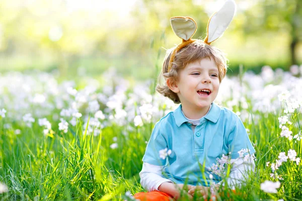 Cute little kid boy with Easter bunny ears celebrating traditional feast. Happy child smiling on warm sunny day. Family, holiday, spring concept. Toddler sitting on grass between flowers. — Stock Photo, Image