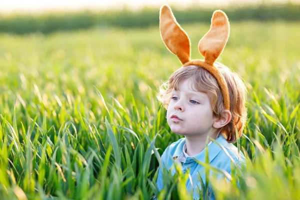 Cute little kid boy with bunny ears having fun with traditional Easter eggs hunt on warm sunny day, outdoors. Celebrating Easter holiday. Toddler finding, colorful eggs in green grass — Stock Photo, Image