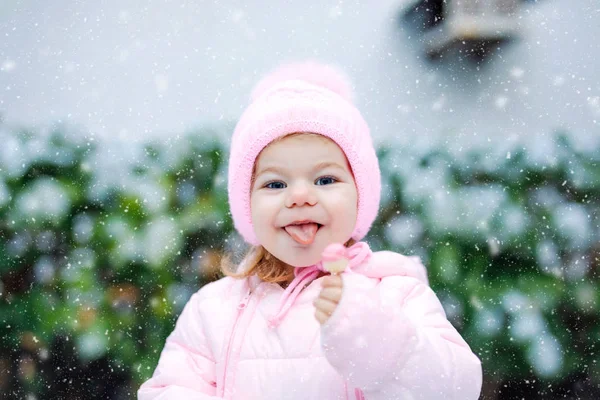 Portrait de petite fille marchant à l'extérieur en hiver. Bébé mignon mangeant des bonbons sucrés sucrés. Enfant qui s'amuse par temps froid. Porter des vêtements chauds bébé rose et chapeau avec des bobbles. — Photo