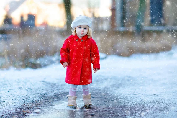 Utomhus vintern porträtt av små söta småbarn flicka i röd rock och vitt mode hatt barret. Friska glada barn promenader i parken på kall dag med snö och snöfall. Snygga kläder för barn. — Stockfoto