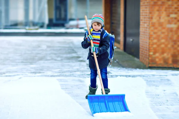 Маленький школяр хлопчик початкового класу прибирає сніг лопатою, перш ніж піти до школи. Щаслива дитина розважається з першим снігом. Студент з рюкзаком або сателітом в барвистому зимовому одязі . — стокове фото