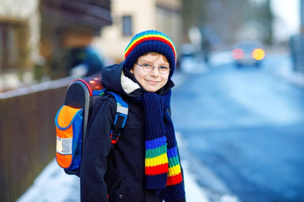 Kleine schooljongen van de lagere klasse die naar school loopt tijdens de sneeuwval. Gelukkig gezond kind met een bril die plezier heeft en met sneeuw speelt. Met rugzak of tas in kleurrijke winterkleding. — Stockfoto