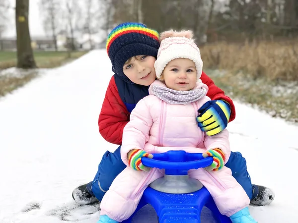 Mały chłopiec i urocza dziewczynka siedzą razem na saniach. Rodzeństwo, brat i młodsza siostra cieszą się jazdą saniami podczas opadów śniegu. Dzieci jeżdżące na śniegu. Aktywna zabawa na rodzinne wakacje — Zdjęcie stockowe