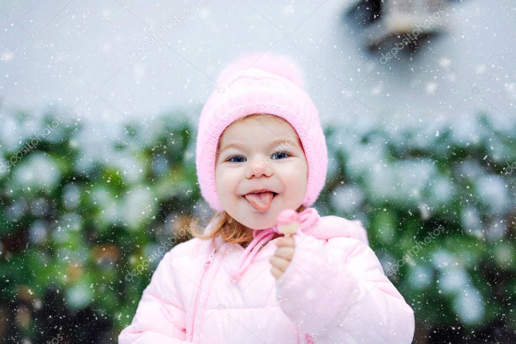 Portrait of little toddler girl walking outdoors in winter. Cute toddler eating sweet lollypop candy. Child having fun on cold snow day. Wearing warm baby pink clothes and hat with bobbles.