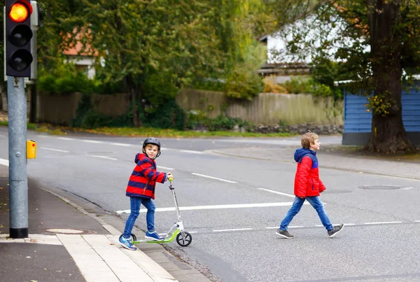 Çalışan ve sonbahar gününde scooter üzerinde sürüş iki küçük schoolkids çocuklar. Renkli giysiler ve şehir trafik geçerken yaya yaya geçidi trafik ışıkları ile mutlu çocuk. — Stok fotoğraf