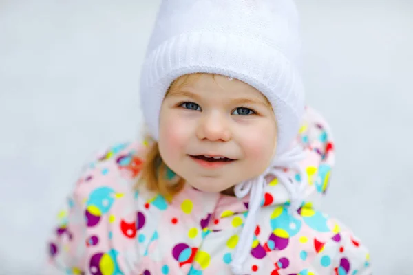 Portret van een klein peutermeisje dat 's winters buiten loopt. Leuke peuter die zoete lolly snoep eet. Kind heeft plezier op koude sneeuwdag. Het dragen van warme baby kleurrijke kleding en hoed met bobbels. — Stockfoto