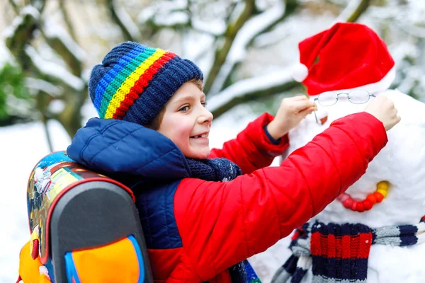 Little school kid boy in colorful clothes, with glasses and backpack having fun with snowman after elementary school end — Stock Photo, Image