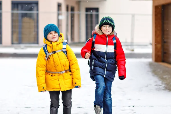 Two little kids boys of elementary class walking to school during snowfall. Happy children having fun and playing with first snow. Siblings and best friends with backpack in colorful winter clothes. — Stock Photo, Image