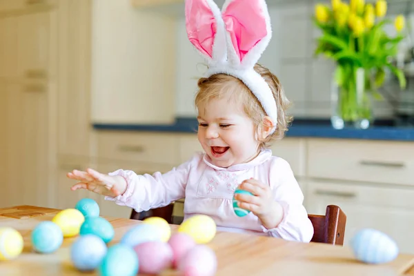 Милая маленькая девочка с пасхальными кроличьими ушами, играющая с цветными пастельными яйцами. Счастливый ребенок распаковывает подарки. Очаровательный ребенок в розовой одежде наслаждается праздником Стоковая Картинка