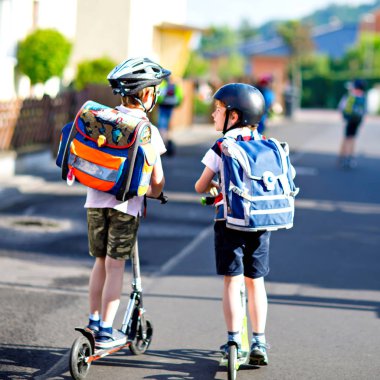 Güneşli bir günde, ellerinde scooterla şehirde gezen kasklı iki okul çocuğu. Renkli giysiler içinde okula bisikletle giden mutlu çocuklar..