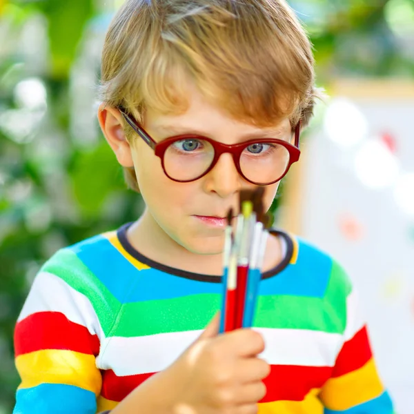 Menino confuso com óculos segurando aquarelas e pincéis. Criança feliz e estudante está de volta à escola. Educação, escola, conceito de aprendizagem. Escola, pré-escolar equipamentos de berçário . — Fotografia de Stock