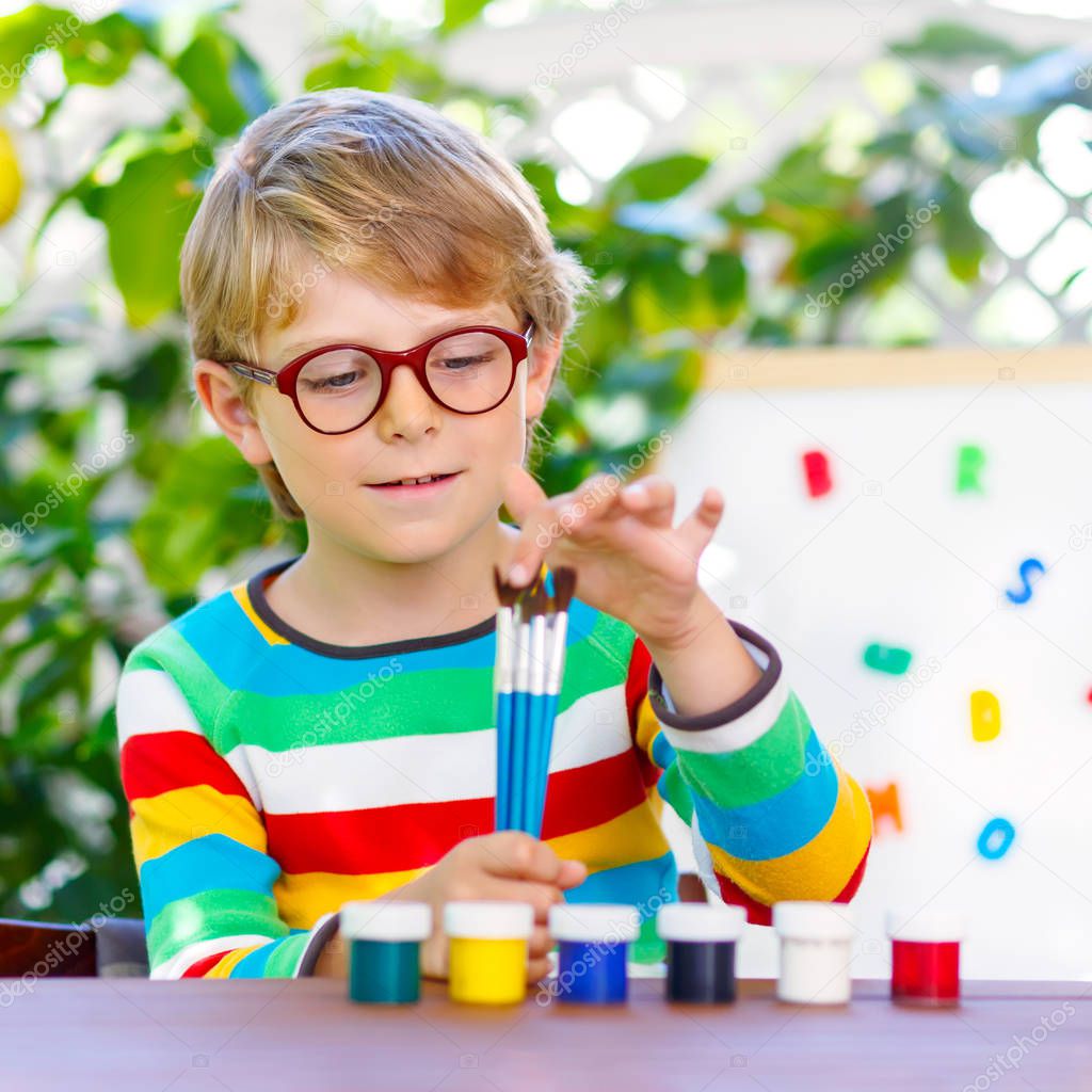 Funny adorable little kid boy with glasses holding watercolors and brushes. Happy child and student is back to school. Education, school, learning concept. School, preschool nursery equipment.