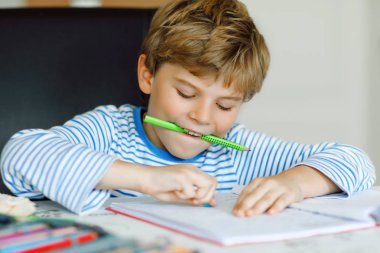 Evde ev ödevi yapan sağlıklı, şirin bir okul çocuğunun portresi. Renkli kalemlerle, kapalı alanda yazan küçük bir çocuk. İlkokul ve eğitim. Çocuk harf ve sayı yazmayı öğreniyor