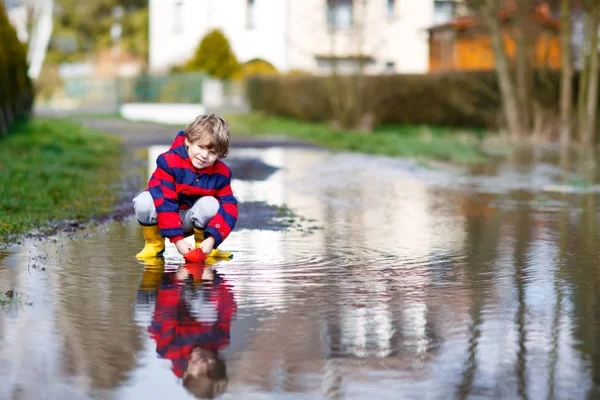 Menino pequeno feliz em botas de chuva amarela jogando com barco de navio de papel por enorme poça no dia de primavera ou outono. Lazer ativo para crianças. Criança engraçada se divertindo ao ar livre, vestindo roupas coloridas. — Fotografia de Stock