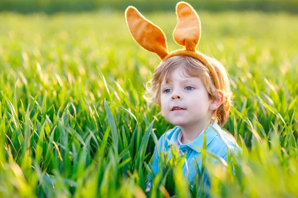 Cute little kid boy with bunny ears having fun with traditional Easter eggs hunt on warm sunny day, outdoors. Celebrating Easter holiday. Toddler finding, colorful eggs in green grass — Stock Photo, Image