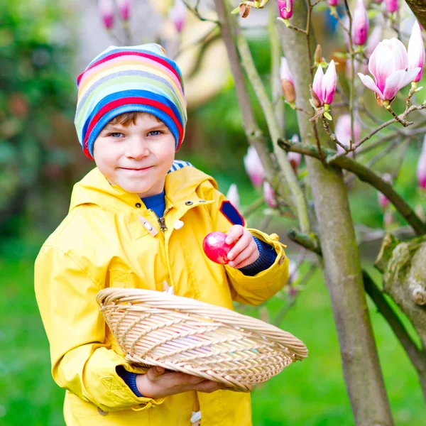 Cute adorable little kid boy making an egg hunt on Easter. Happy child searching and finding colorful eggs in domestic garden. Boy in spring clothes on cold day. Old christian and catholoc tradition. — Stock Photo, Image