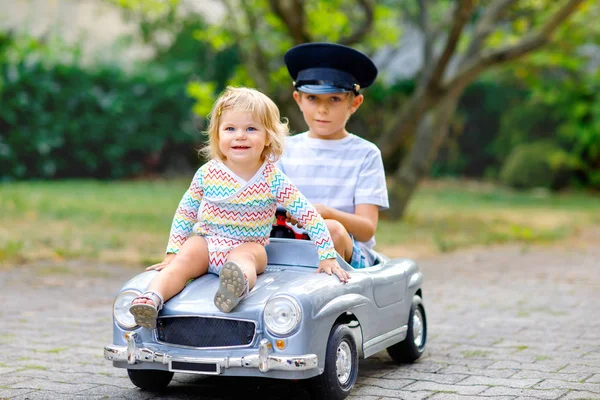 Duas crianças felizes brincando com o carro de brinquedo velho grande no jardim de verão, ao ar livre. Rapaz carro de condução com a menina da criança lá dentro. Miúdos risonhos e sorridentes. Família, infância, conceito de estilo de vida — Fotografia de Stock
