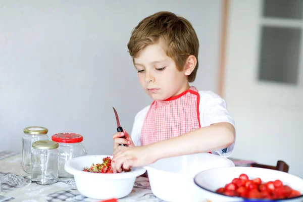 Niño rubio ayudando y haciendo mermelada de fresa en verano. Funny niño limpiando bayas y preparándose para la mermelada de cocina. Niño comiendo fresas maduras en la cocina doméstica.. —  Fotos de Stock