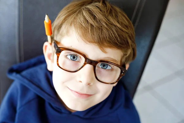 Ritratto di un ragazzino carino con gli occhiali. Bella bambina felice che guarda la macchina fotografica. Schoolboy prendere in giro con una penna, tenendo in bocca e dietro l'orecchio. Concetto educativo. — Foto Stock