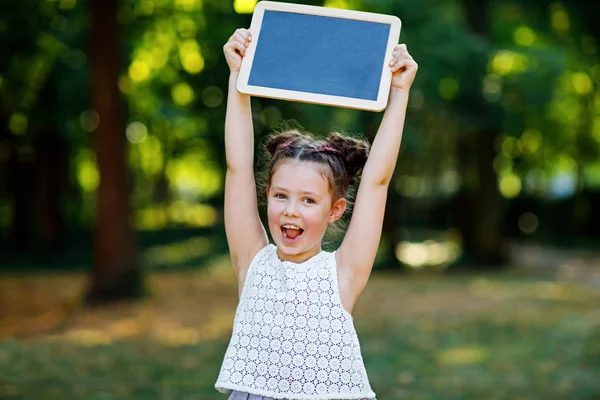 Happy little kid girl holding empty chalk desk in hands. Schoolkid on first day of elementary class. Healthy adorable child outdoors, in green park. Copyspace on desk — Stock Photo, Image