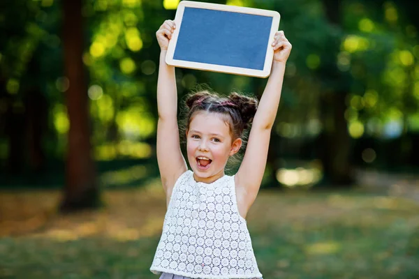 Ragazzina felice che tiene in mano una scrivania vuota di gesso. Studente il primo giorno di lezione elementare. Bambino sano adorabile all'aperto, nel parco verde. copyspace sulla scrivania — Foto Stock