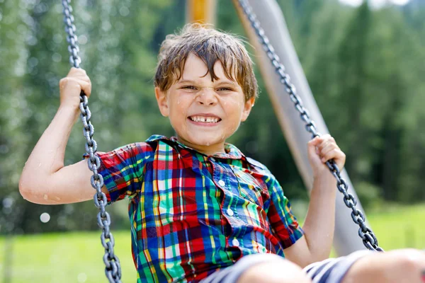 Zabawny chłopiec bawiący się huśtawką łańcuchową na placu zabaw na świeżym powietrzu, będąc mokrym rozbryzganym wodą. kołyszące się dziecko w letni dzień. Aktywny wypoczynek z dziećmi. Szczęśliwy płaczący chłopiec z kroplami deszczu na twarzy. — Zdjęcie stockowe