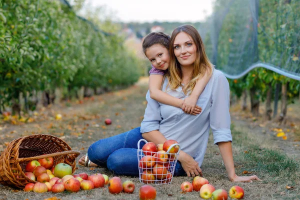 Portrait of little girl and beautiful mother with red apples in organic orchard. Happy woman and kid daughter picking ripe fruits from trees and having fun. Harvest season for family. — Stock Photo, Image