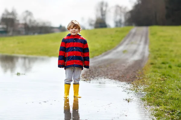 Little kid boy wearing yellow rain boots and walking and jumping into puddle on warm sunny spring day. Happy child in colorful fashion casual rain clothes having fun and playing outdoors. — Stock Photo, Image