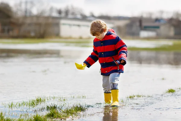 Little kid boy wearing yellow rain boots and walking and jumping into puddle on warm sunny spring day. Happy child in colorful fashion casual rain clothes having fun and playing outdoors. — Stock Photo, Image