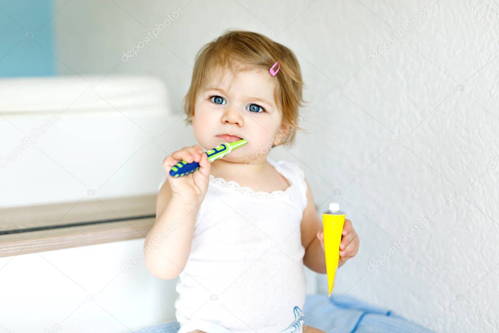 Little baby girl holding toothbrush and brushing first teeth. Toddler learning to clean milk tooth. Prevention, hygiene and healthcare concept. Happy child in bathroom