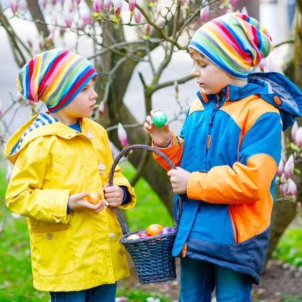 Duas crianças meninos e amigos fazendo tradicional caça ao ovo de Páscoa no jardim da primavera, ao ar livre. Irmãos se divertindo com a descoberta de ovos coloridos. No dia frio. Velha tradição cristã e católica . — Fotografia de Stock