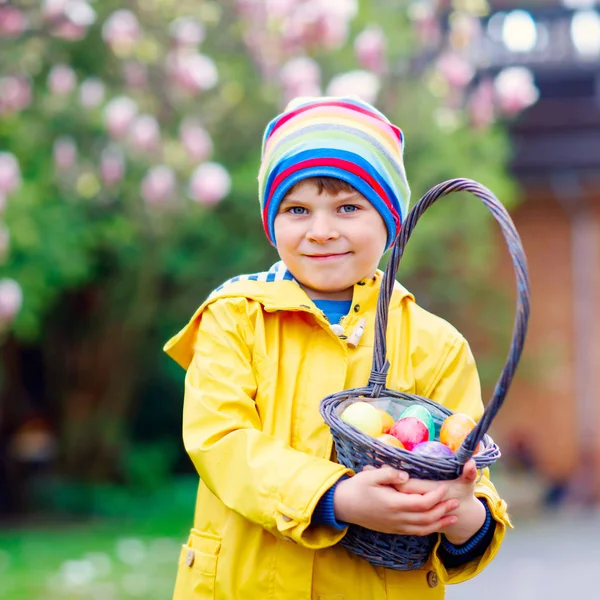 Carino adorabile ragazzino che fa una caccia alle uova a Pasqua. Bambino felice che cerca e trova uova colorate nel giardino domestico. Ragazzo in abiti primaverili nelle giornate fredde. Vecchia tradizione cristiana e cattolica . — Foto Stock