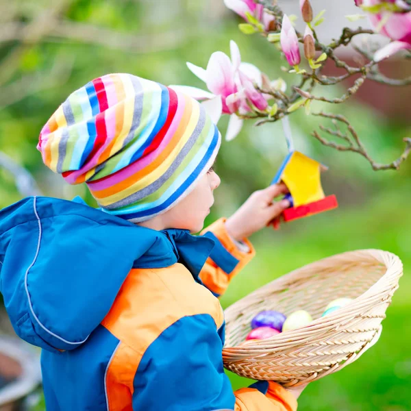 Schattige schattige kleine jongen jongen maken een ei jagen op Pasen. Gelukkig kind zoeken en vinden van kleurrijke eieren in binnenlandse tuin. Jongen in voorjaar kleren op koude dag. Oude christelijke en katholieke traditie. — Stockfoto