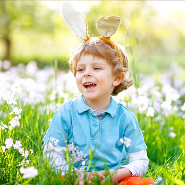 Sevimli küçük çocuk çocuk Paskalya tavşanı ile kutluyor geleneksel bayram kulaklar. Sıcak güneşli günde gülümseyerek mutlu bir çocuk. Aile, tatil, Bahar kavramı. Çimen çiçekler arasında oturan yürümeye başlayan çocuk.