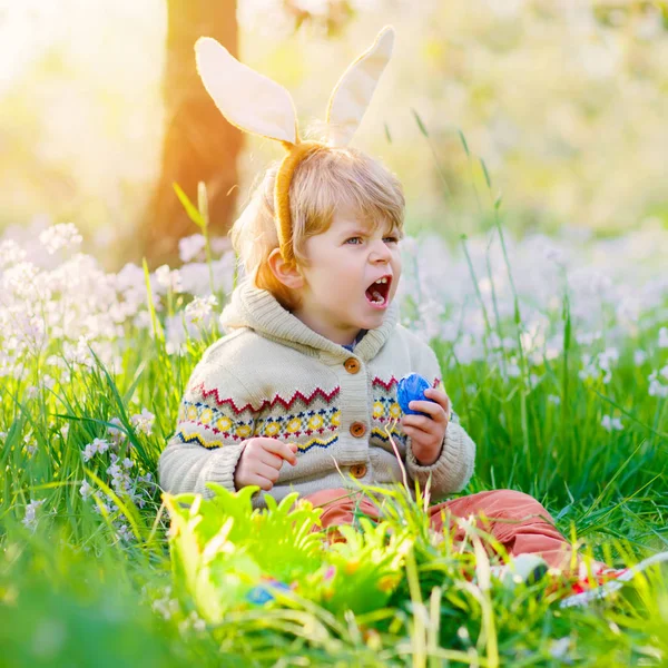 暖かい晴れた日に、屋外で伝統的なイースターエッグハントを楽しんでいるウサギの耳を持つかわいい子供の男の子。イースターの祝日を祝う。幼児の発見,緑の芝生の中でカラフルな卵 — ストック写真
