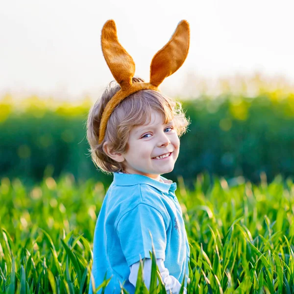 Cute little kid boy with bunny ears having fun with traditional Easter eggs hunt on warm sunny day, outdoors. Celebrating Easter holiday. Toddler finding, colorful eggs in green grass — Stock Photo, Image