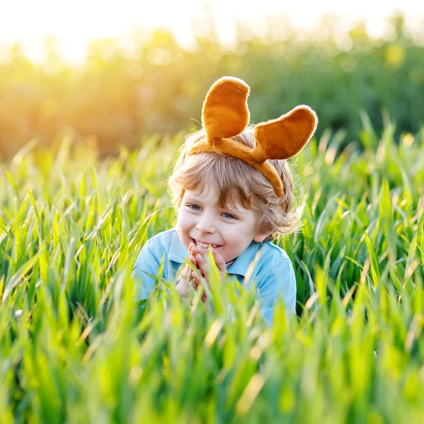 Petit garçon mignon aux oreilles de lapin qui s'amuse avec les œufs de Pâques traditionnels chassent par une chaude journée ensoleillée, en plein air. Fête de Pâques. Recherche de tout-petits, oeufs colorés dans l'herbe verte — Photo