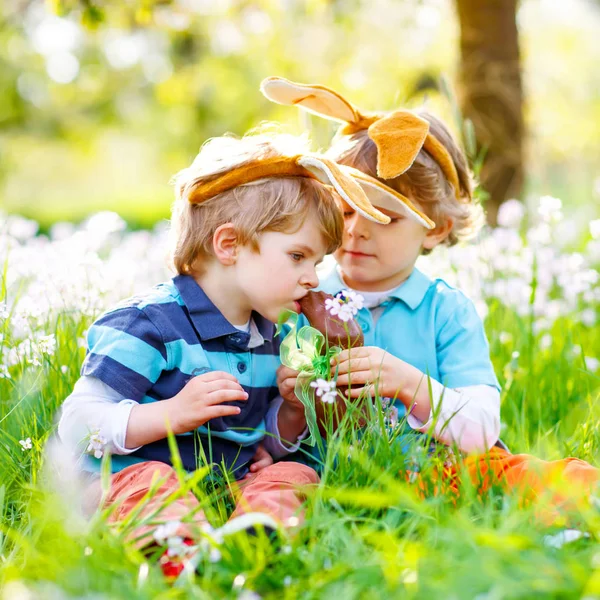 Two little kids boys and friends in Easter bunny ears during traditional egg hunt in spring garden, outdoors. Siblings eating chocolate bunnies and eggs. Old christian and catholoc tradition. — Stock Photo, Image