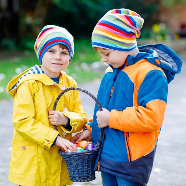 Two little kids boys and friends making traditional Easter egg hunt in spring garden, outdoors. Siblings having fun with finding colorful eggs. On cold day. Old christian and catholoc tradition. — Stock Photo, Image