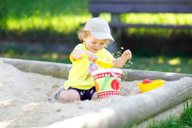 Dışarıda parkta kumlarda oynayan tatlı bir kız çocuğu. Güneşli sıcak yaz gününde güzel bir bebek eğleniyor. Kum oyuncakları ve renkli kıyafetlerle mutlu sağlıklı bir çocuk..