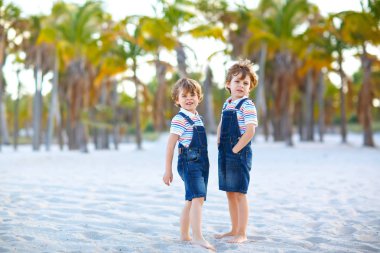 İki küçük çocuk tropikal plajda eğleniyor, mutlu arkadaşlar oynuyor, arkadaşlık kavramı. Kardeşler, ailede ikizler arka planda palmiye ağaçları olan tipler. Aile tatilleri.